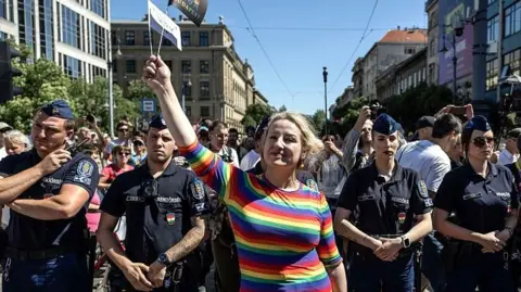 Getty Images A woman in a rainbow coloured top defiantly waves a flag as she stands in front of a row of police officers at Budapest Pride.