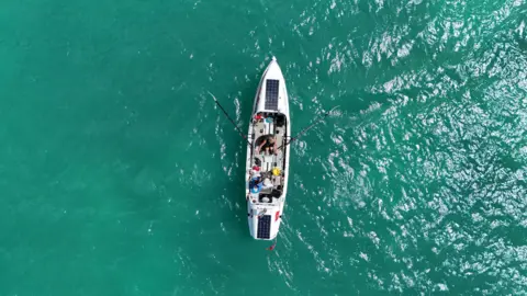 ATLANTIC DASH An arial view of the boat, Mike is rowing, and Tim is stood up, it is a small boat with solar panels, and thy are in turquoise waters.