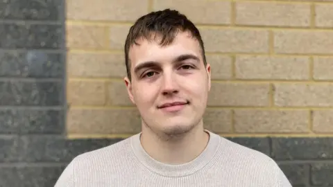 A man, with brown hair, standing in front of a brick wall and looking at the camera. He is wearing a cream coloured jumper. 