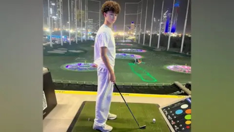 Submitted Noah stands side-on with a golf club in his hand at a golf simulator. Skyscrapers can be seen in the distance. He wears a white T-shirt, joggers and trainers. The picture has been taken at night.