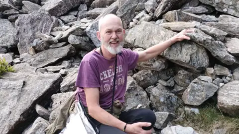 Family Handout A bearded middle-aged man kneeling by some rocks whilst wearing a purple shirt with obscured writing on it.