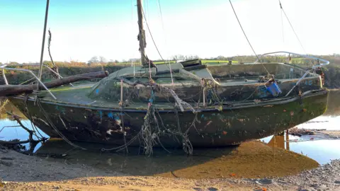 A small yacht on a river bed covered with debris including a tree trunk, weed and fungus.