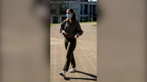 A student wearing a face mask walks on the University of Kent campus. She is wearing dark green trousers and a brown jacket.