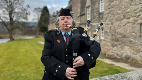 BBC Seorang wanita yang mengenakan gaun formal dataran tinggi memainkan bagpipe. Dia memiliki rambut abu -abu dan mengenakan jaket hitam. Ada bangunan tua dan pohon di latar belakang.