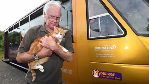 West Midlands Railway An orange metro tram with "george the station cat" purple and orange sticker on it. Infront of it is a man with white short hair in glasses looking serious as he holds orange tabby George in his arms. 