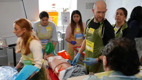 A medical training exercise. Several people wearing surgical gloves and aprons are working on a dummy patient.