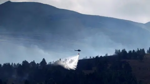 Getty Images Water pours from a large container slung beneath a helicopter over an area of moor and forestry. The helicopter is silhouetted against a mountainside. 