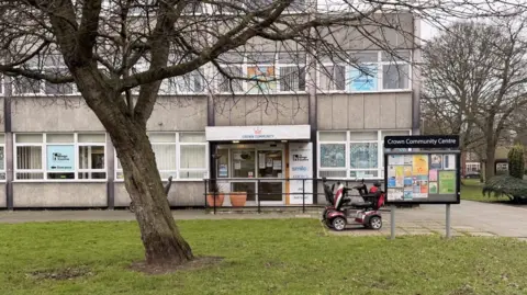 The exterior of the Crown Community Centre offices. A large tree is in front of the three-storey building. A mobility scooter is parked behind a notice board outside of the building. 