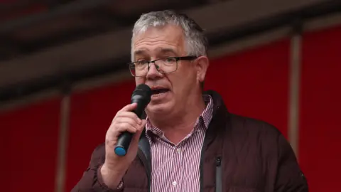 Liam McBurney/PA Wire James Geoghegan is on a stage. he's speaking into a mic. He wears glasses and has a stripped shirt on with a maroon coat. 