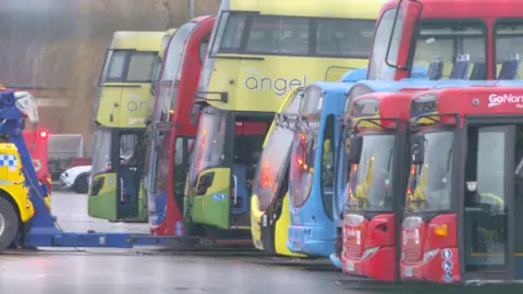 A recovery truck lifts a yellow and green double decker bus off the ground, It is surrounded in the middle of a line of brightly coloured buses.