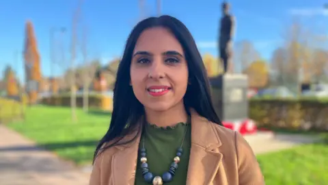 Sandy Kaur has long dark hair and is wearing a brown jacket with a green top and necklace. The Sikh soldier statue can be seen behind her out of focus.