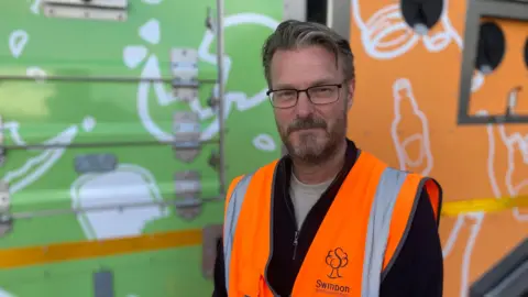 Councillor Chris Watts - in glasses, with a beard and wearing orange high-vis - looks into the camera in front of a recycling lorry.