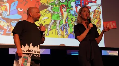 La Vida Liverpool & Jade Andreas A man stands next to a woman on a stage. They are both holding microphones and she is speaking into hers and holding a book called Tiny Daggers in her other hand. 
