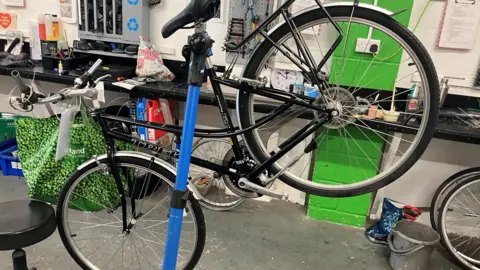 Chris Goreham/BBC A bike suspended in the air so it can be repaired. The front wheel of the bike is on the ground while the back of the bike and the back wheel are in the air. It is being propped up by a blue pole. The bike is inside a workshop.