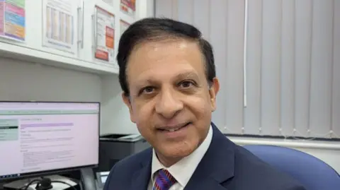 Dr Chaand Nagpaul is pictured looking straight into the camera, smiling. He is sat on a blue chair with white blinds in the background and a computer screen showing a health website. 