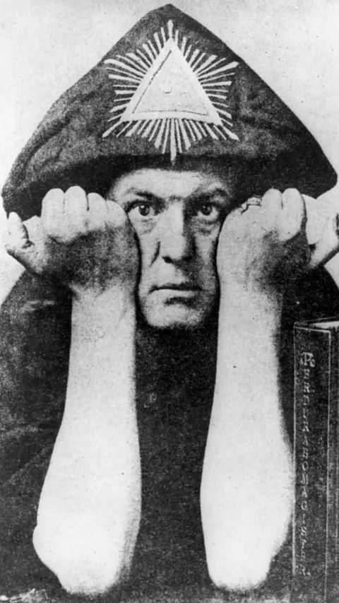 Getty Images Black-and-white portrait of Aleister Crowley who is seen in a hooded robe marked with a radiant triangle symbol, staring directly at the camera with his fists raised on either side of his face.
