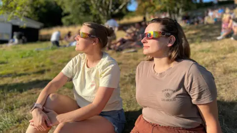 Two women are sat down wearing special eclipse glasses to look at the sun
