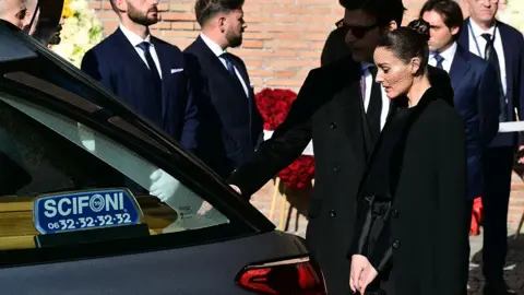 Stefano RELLANDINI / AFP via Getty Images Olivia Palermo dressed in black stands beside a hearse, looking towards the flower-covered coffin as mourners gather nearby.