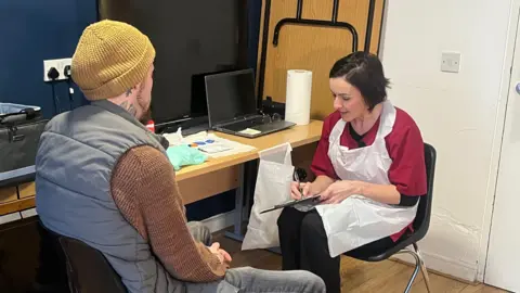 Lisa Ewart is sitting on a chair writing on a clipboard. She has short dark hair, wearing dark red scrubs and a disposable apron. Curtis Durose is sitting opposite her. He is wearing a yellow beanie hat, brown jumper, blue puffer vest and jeans. 