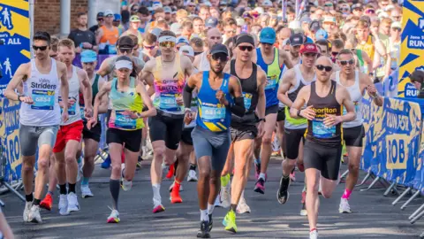 Image shows the start of the Rob Burrow Leeds Marathon with runners in a mixture of coloured and branded running vests on. The street is full of runners and on the side of the road there are signs that read 'RUN FOR ROB'.