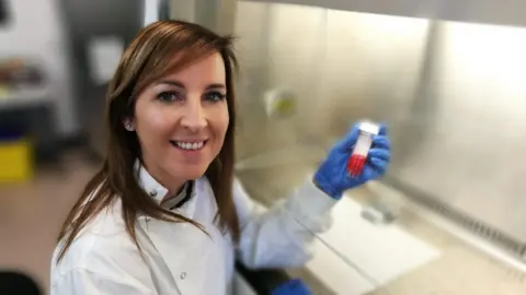 Trisha McCluney Trisha sitting smiling at the camera. She is wearing a white lab coat and blue gloves and is holding a vial with a red liquid in it.