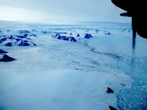 British Antarctic Survey Three glaciers flowing out into the ocean from between mountain ranges. The ocean surface is a mix of open water, icebergs and sea-ice. Part of the aircraft is seen in the top right corner of the image.