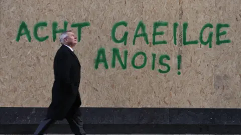 PA media wearing a tall black coat is a man beyond a large corkboard that reads 'Acht Gelz Aneois' in green spray paint.