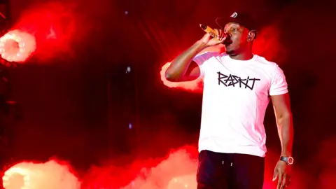 PA Media Dizzee Rascal is on stage at a live gig. He is wearing a white T-shirt and a baseball cap