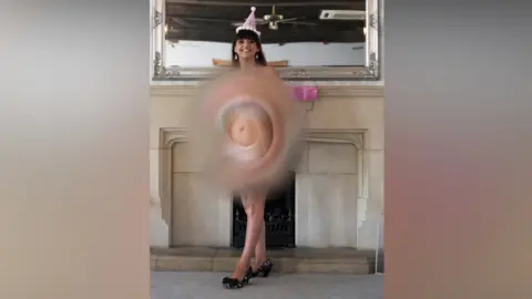 Deafblind UK A woman is standing in front of a front of a fireplace and a mirror smiling, wearing a pink hat and is naked.