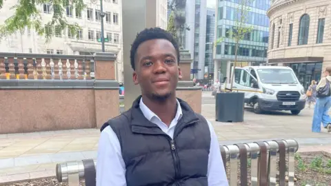 Erin McLone/BBC Sat on a metal bench in Leeds City Square, a man looks and smiles at the camera. He wears a black gilet over a light blue dress shirt.