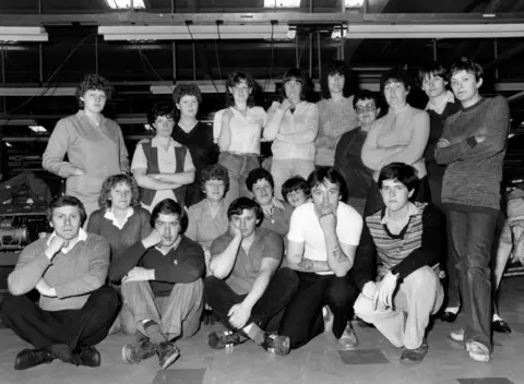 Mirrorpix via Getty Images A black and white shot of factory workers in the 1980s. Men and women are pictured in three rows, those at the back are standing and those at the front are sitting.
