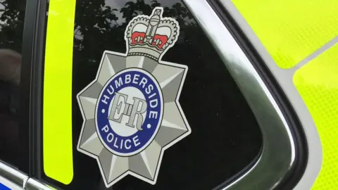 Humberside Police A Humberside Police logo on the side of a police car. It is a silver star with a crown at the top with ER and Humberside Police in a circle in the middle. You can see the yellow high vis on the police car.