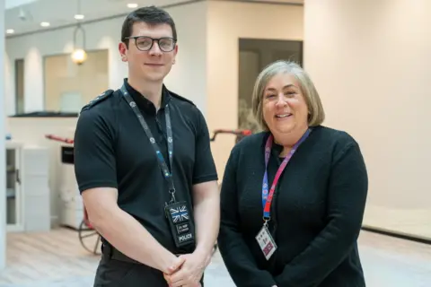 Police, Fire and Crime Commissioner’s Office A man with a black lanyard, black t-shirt and dark glasses. He has dark hair, he is smiling at the camera and his left handhis holding his right arm. A woman with a black jumper and red and blue lanyard is smiling at the camera. Her hair is in bob.