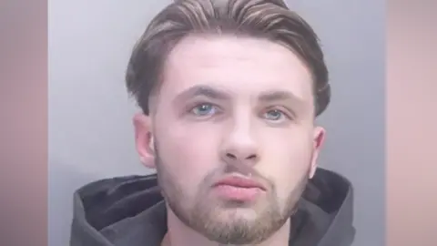 Police mugshot of Chas Corrigan. He is wearing a dark hoodie and has brown hair swept back and a beard.