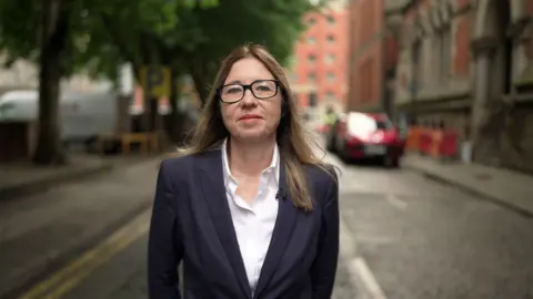 Brijesh Patel / BBC Rachel is pictured dressed smartly with long hair, wearing a dark blazer over a white shirt. She is wearing glasses and stands on a city street. The background shows trees, buildings, and parked vehicles.