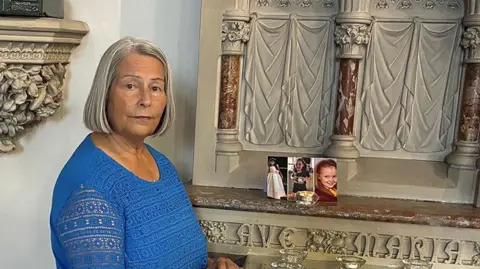 Sacristan Evelyn Hart with a blond bob wearing a royal blue patterned top stands at a church altar with the images of the three young girls - Alice da Silva Aguiar, Bebe King and Elsie Dot Stancombe who died in the Southport knife attacks - with a candle.