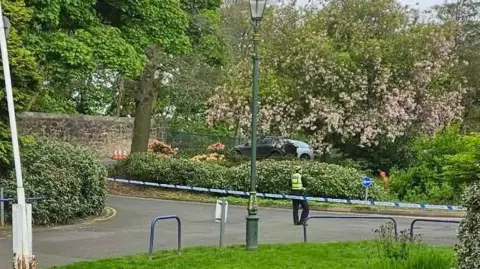 Police tape cuts off the area of a park with green grass and pink blossom trees