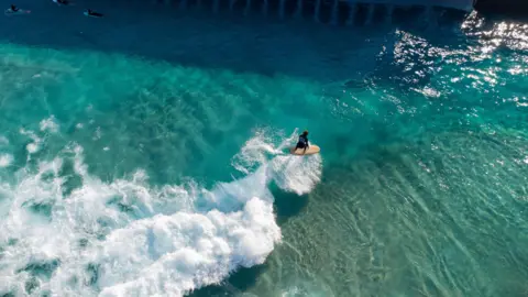 Image Kabin An aerial picture of a surfer riding a white water wave. The water is clear and bright blue, and is sparkling in the sun.