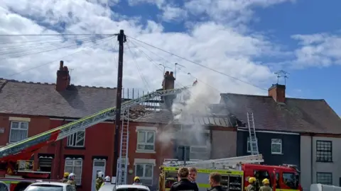 Sara Bibb A row of terraced houses where the roof of the middle house is fire damaged. There is a fire engine in front of the house and a crane pouring water onto the damaged roof. Fire crews are gathered in front of the house.