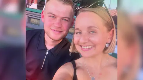 Charlotte Vockins A selfie of Charlotte and her partner Ryan smiling at the camera. She has blue eyes, a short blonde bob and is wearing sunglasses on her head and silver heart earrings. Ryan has short brown hair and green-grey eyes, and is wearing a black three-quarter zip shirt.