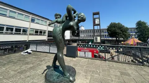 Ben Schofield/BBC Paved stage area in Stevenage town centre with a bronze sculpture of a mother carrying a child (known as "Joyride"). There are black railings around the stage. Three-storey shop and office buildings are visible behind, along with two trees.