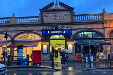 BBC/Harry Low Exterior of Barons Court station shows underground sign and woman with umbrella 