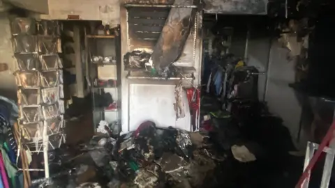 The Children's Society A badly burnt shop with lots of damaged goods on the floor. There is CD rack on the left, a burned material hanging from the ceiling and racks of clothes to the right.