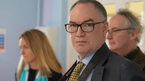 John Clark is stood in front of two people with a Canon camera strap around his neck, wearing a blue shirt and a dark suit. He has a yellow tie.
