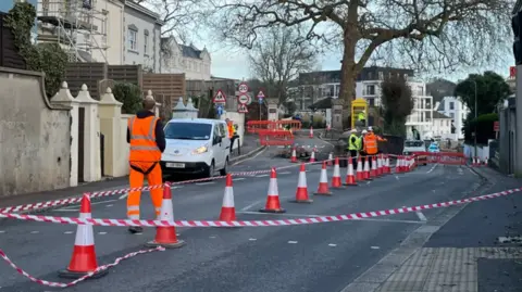 A picture of the scene where the sinkhole is. Cones can be seen separating a section of the road off. There are workers walking around with tape.