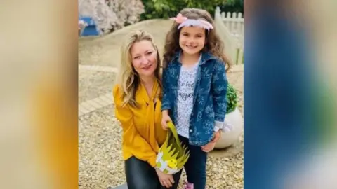 Zoe Hoes, una mujer que tiene cabello rubio con una tapa amarilla. Ella está sosteniendo a una niña alrededor de su cintura mientras la pareja sonríe para una foto. La niña tiene pintura en la cara de gato en su rostro y lleva una chaqueta de mezclilla azul y una blusa blanca. Están parados en un área de piedra.