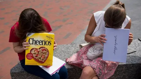 Two girls look into cardboard boxes, one a cereal box and another a shoe box, while sitting on a bench outside.