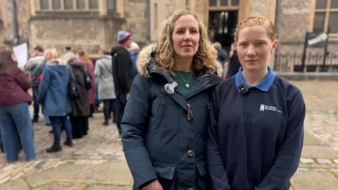 A mum and daughter - the girl has ginger hair and is in her navy school uniform with the Henry Beaufort logo on her jumper. The mum is in a navy coat. They are both stood outside the registry office behind the group of protesters.