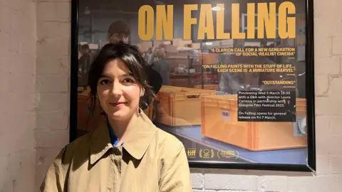 Film maker Laura Carreira stands in front of a poster of her film 'On Falling'. The poster features a woman stacking a yellow box onto a conveyer belt in a warehouse. Some of the reviews read "A clarion call for a new generation of social-realist cinema" and "Outstanding".