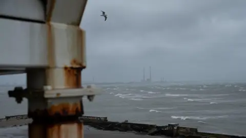Getty Images A dark, grey sky. A seagull is flying over the sea. A metal pillar is in the foreground. Visibility is poor. 